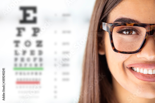 Close up of young woman wearing eyeglasses with eyechart in the background. Eyewear. Closeup view of young woman and blurred eye chart on background. Space for text photo