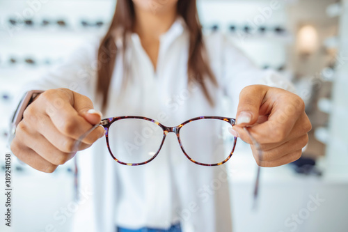 Optician showing and suggesting eyeglasses in optical shop. Cheerful female ophthalmologist is working with patient. Optician giving new glasses to customer for testing and trying.