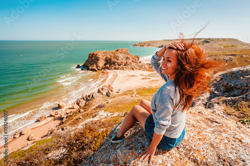 A happy woman with long hair smiles and tilts her head back as she sits on the rocks overlooking the rocky ocean shore