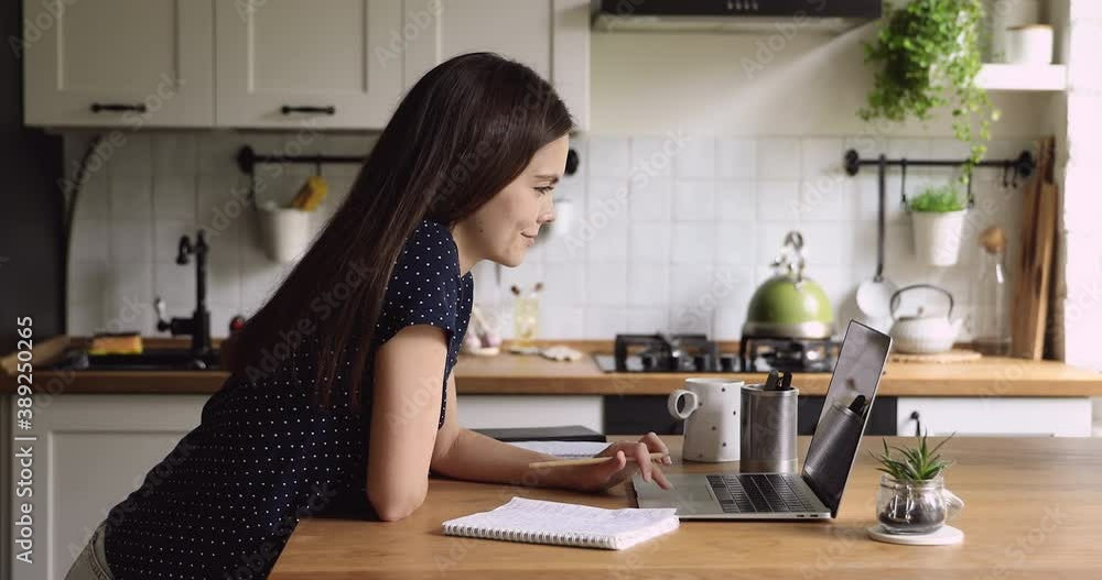Side view pleasant young woman leaning on table, web surfing information on computer, handwriting recipes in kitchen. Smiling 20s female student studying remotely on educational courses at home. Side view pleasant young woman leaning on table, web surfing information on computer, handwriting recipes in kitchen. Smiling 20s female student studying remotely on educational courses at home.