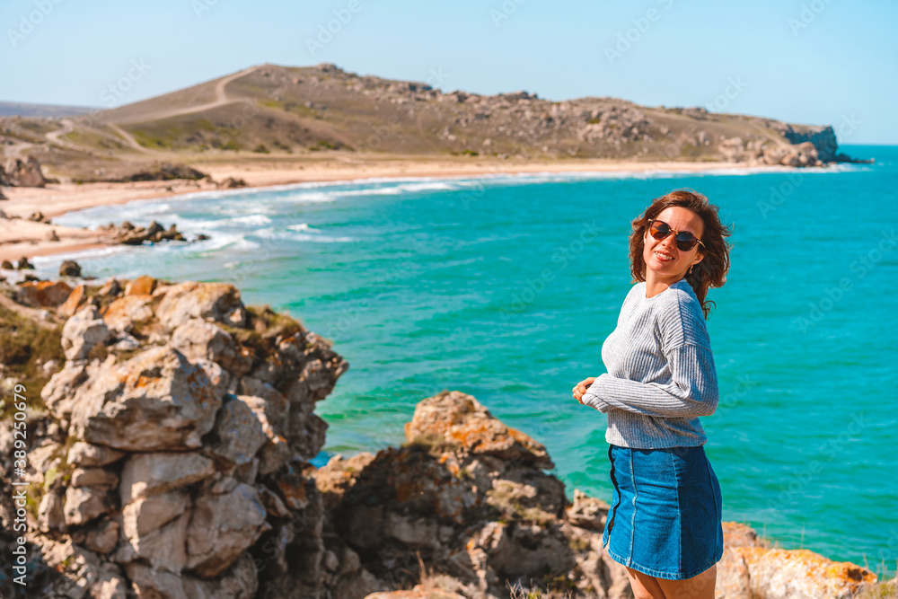 A happy woman with long hair sits on a rock with her back to the camera and admires the raging sea in the middle of the rocks