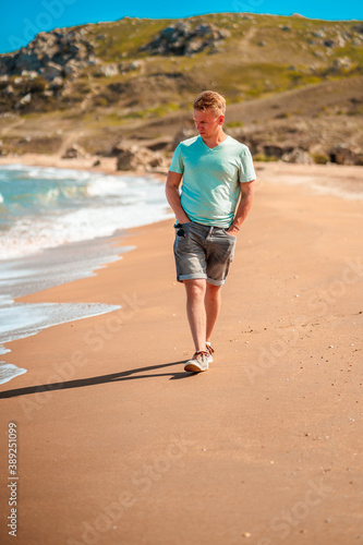 Handsome young blond man walks along the Black sea on the beach in sunny day