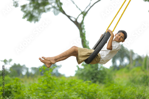 Cute indian child playing on swing made by tyre and rope on tree at green field
