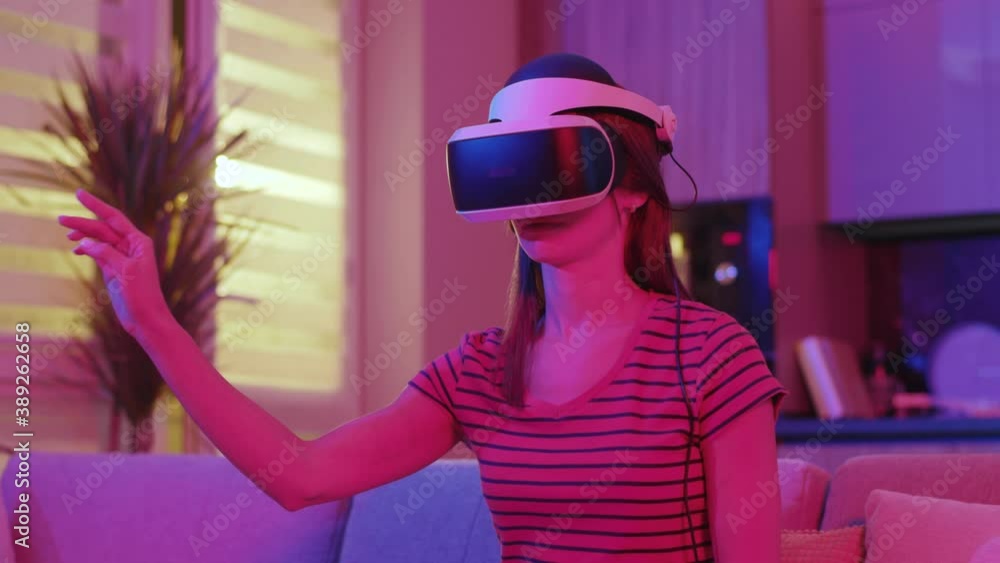 Young woman in VR glasses sitting on sofa at home. In neon light. Moving hands. Technology isolated game innovative. Close up. Slow motion Young woman in VR glasses sitting on sofa at home. In neon light. Moving hands. Technology isolated game innovative. Close up. Slow motion