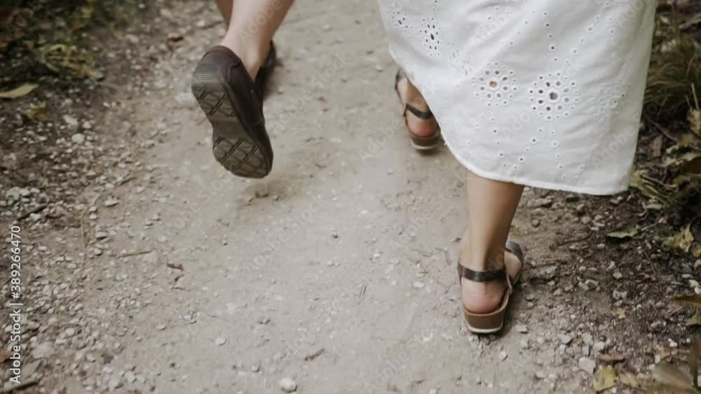 Caucasian couple, man in moccasins and a girl in sandals and a white long dress walk along the path with stones and leaves on both sides. Legs back to knees close up