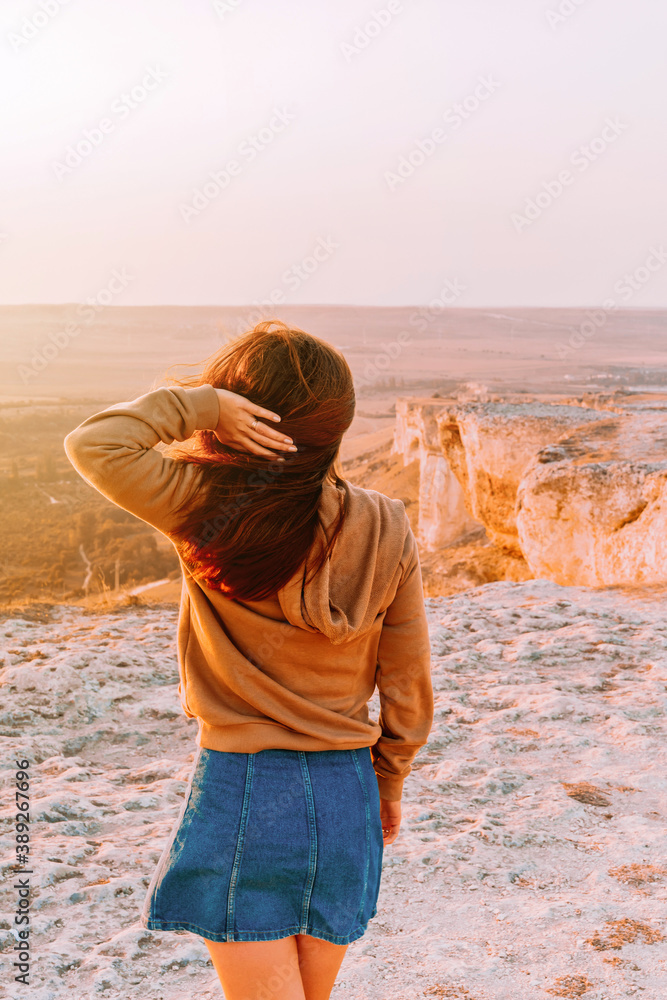 Rear view of a woman holding her long hair and walking on a rocky mountain in an orange sunset, the concept of tourism and freedom