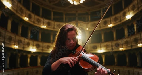 Cinematic shot of professional female violinist is playing violin solo on a classic theatre stage with dramatic lighting before start of a show.