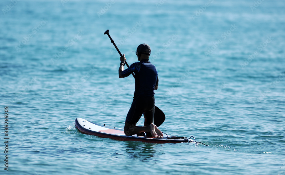 A young man in a wetsuit kneels on his paddle board and paddles.