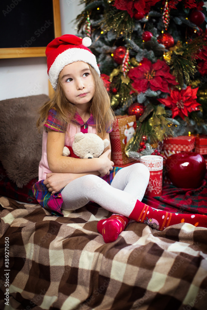 A little girl holding a Teddy bear, sitting on a plaid blanket in the Christmas decorations near a Christmas tree with boxes of gifts and a Santa hat. New year, children's game A little girl holding a Teddy bear, sitting on a plaid blanket in the Christmas decorations near a Christmas tree with boxes of gifts and a Santa hat. New year, children's game