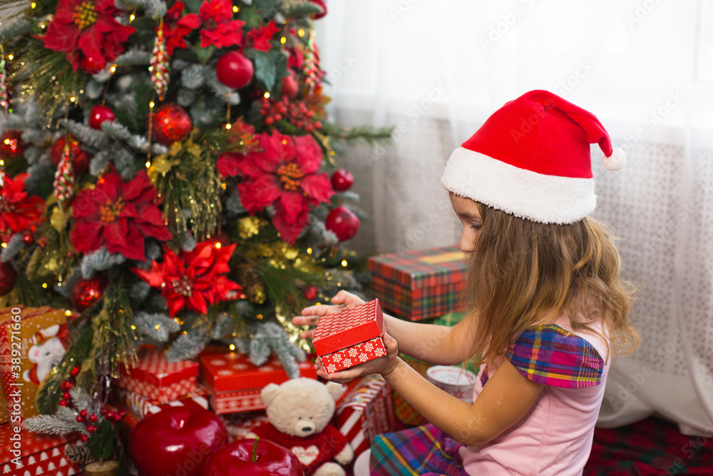 Little girl in Santa hat opens a red box with a gift and a Golden magic light near the Christmas tree. Holiday decor, poinsettias on fir trees, New year. Joy, surprise, children's emotions. Copyspace Little girl in Santa hat opens a red box with a gift and a Golden magic light near the Christmas tree. Holiday decor, poinsettias on fir trees, New year. Joy, surprise, children's emotions. Copyspace