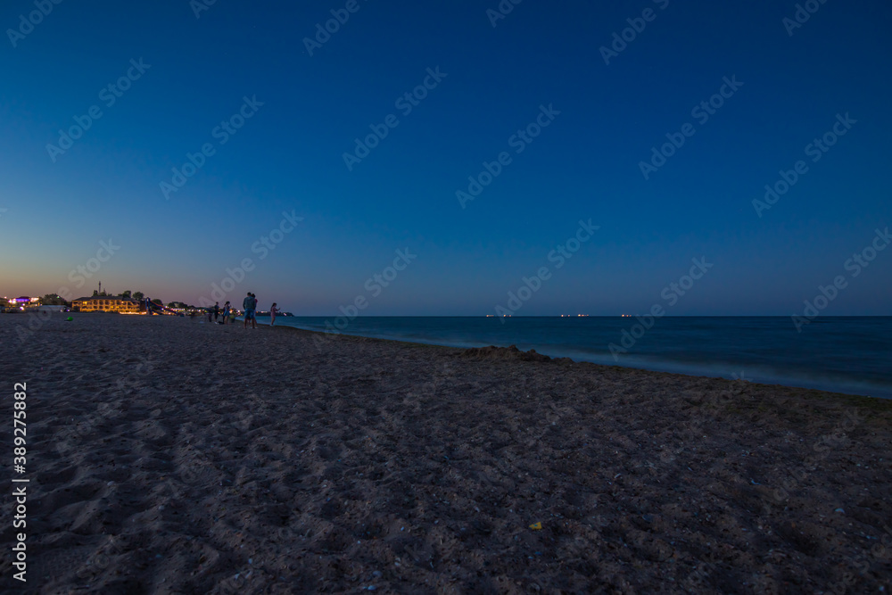 Silhouettes of peoples on the sandy beach after sunset. Blue hour. Lanterns of buildings are visible in the distance