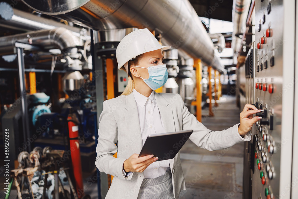 Hardworking independent female supervisor in suit, with helmet and face mask standing next to dashboard and adjusting settings while standing in heating plant and holding tablet during corona virus.