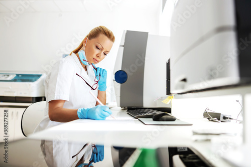Attractive female lab assistant in sterile uniform with rubber gloves on sitting in laboratory and writing down test results.