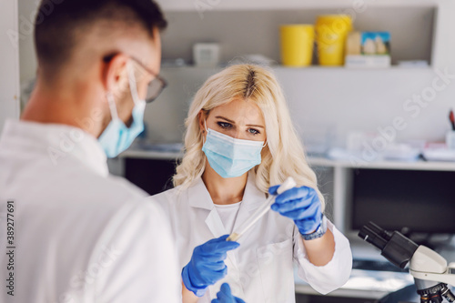 Attractive dedicated blond female lab assistant with face mask and rubber gloves standing in laboratory and holding test tube. She is talking to her colleague about research for the vaccine.