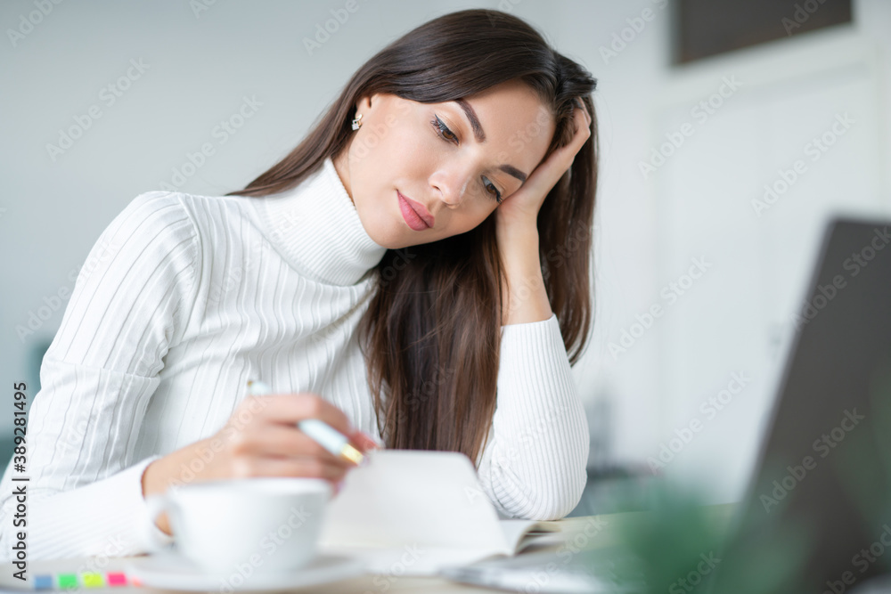 Tired young woman sits desk in front of aptop and turns page of her workbook. Tired young woman sits desk in front of aptop and turns page of her workbook.