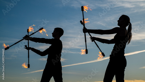 Sexi couple of guy and girl perform flaming baton twirling during fire performance in dark silhouettes on idyllic evening sky outdoors, festival