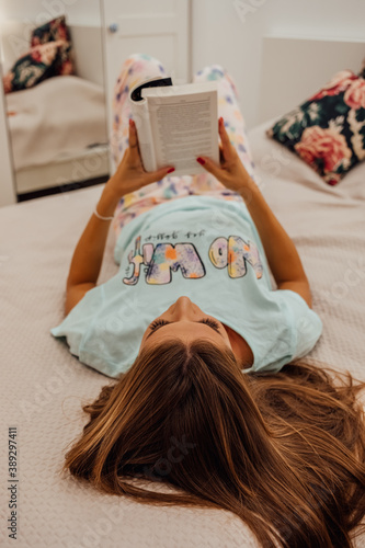 A beautiful young woman in pajamas reading a book while she laying on a bed.