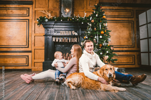 Theme Christmas and New Year family circle. Young Caucasian family with 1 year old child dog breed Labrador Golden Retriever sitting on wooden floor home in living room near fireplace Christmas tree
