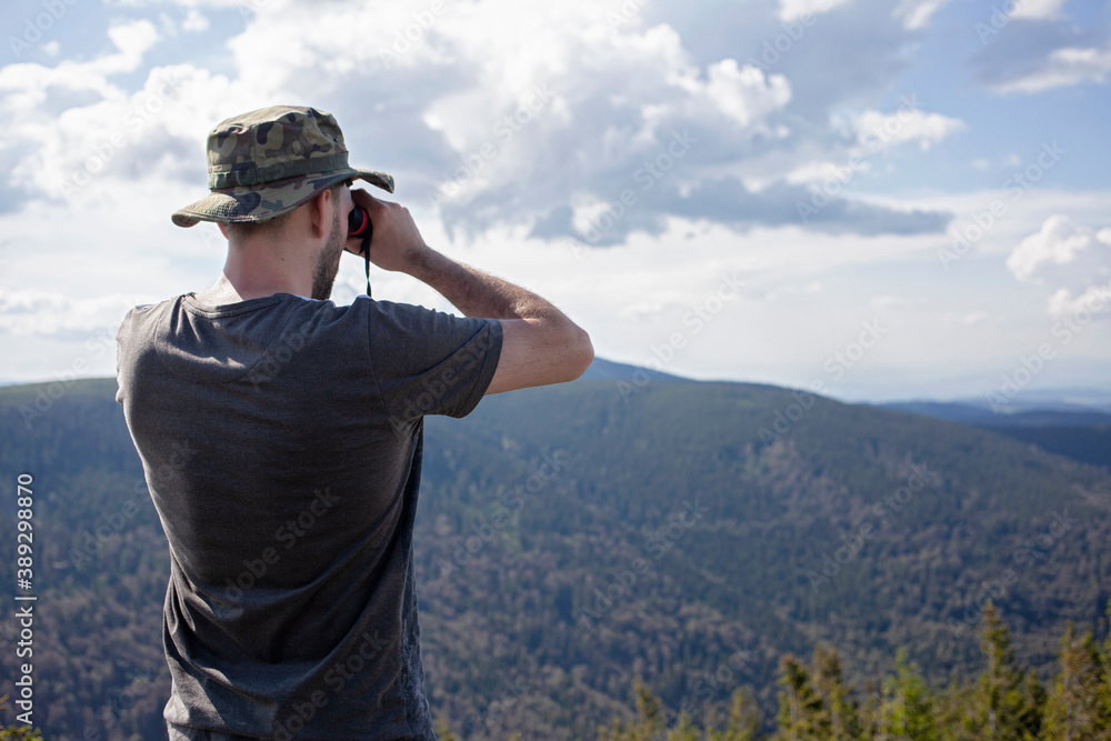 Man looking through binoculars wearing hat and tshirt at mountain hill nature forest landscape. travel adventure observer future concept Man looking through binoculars wearing hat and tshirt at mountain hill nature forest landscape. travel adventure observer future concept