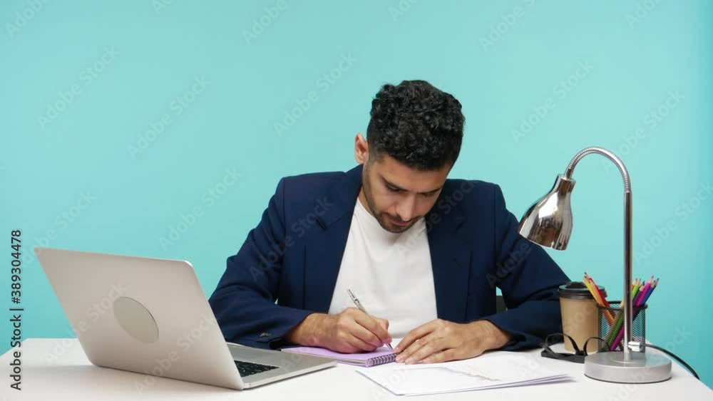 Busy office worker looking at laptop writing down information into notepad, preparing report or to do list, journalist taking notes in office workplace. Indoor studio shot isolated on blue background Busy office worker looking at laptop writing down information into notepad, preparing report or to do list, journalist taking notes in office workplace. Indoor studio shot isolated on blue background