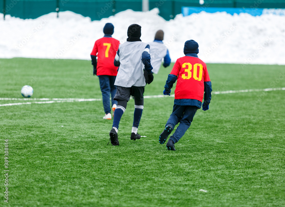 Obraz premium Boys in red sportswear running on soccer field with snow on background. Young footballers dribble and kick football ball in game. Training, active lifestyle, sport, children winter activity