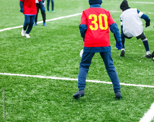 Boys in red sportswear running on soccer field with snow on background. Young footballers dribble and kick football ball in game. Training, active lifestyle, sport, children winter activity