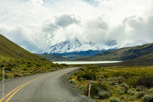 estrada no meio da patagônia, com sua vegetação nativa ao redor, e um lindo lago e montanhas cheias de neve ao fundo