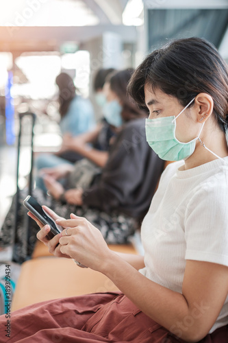 Young female wearing face mask and using mobile smartphone in airport, protection Coronavirus disease (Covid-19) infection, Asian woman traveler sitting on chair. New Normal and social distancing