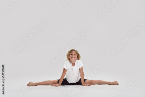 Full length shot of little playful boy child smiling at camera, doing splits isolated over grey background