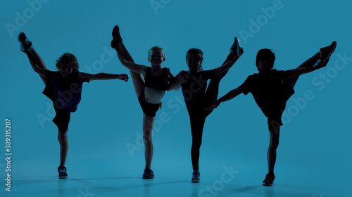 Silhouetted full length shot of four little kids, gymnasts showing flexibility while posing, standing isolated over blue background