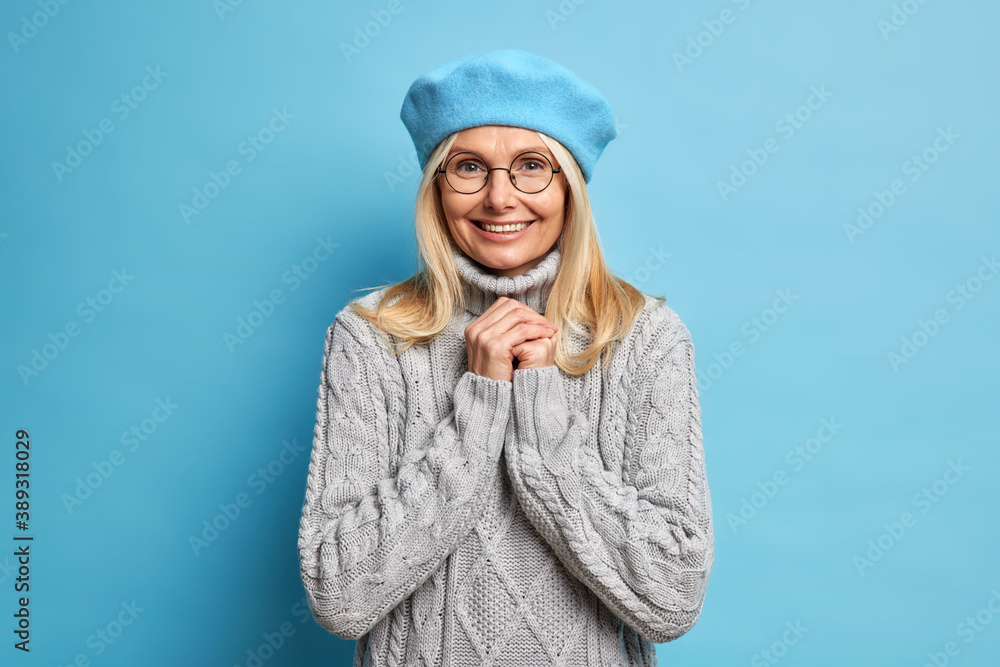 Portrait of good looking adult European woman smiles pleasantly at camera keeps hands together wears optical glasses french beret and knitted sweater expresses positive emotions models in studio