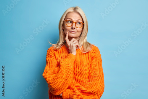 Beautiful middle aged wrinkled woman keeps index finger on cheek stands in thoughtful pose daydreams about something wears knitted sweater isolated over blue background. Need to think deeply photo