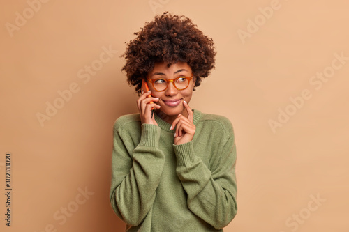 Image of pretty smiling woman talks on mobile phone with friend discuss plans for weekends concentrated aside happily has smart look wears transparent eyewear casual sweater. Technology lifestyle