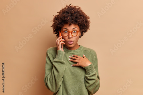 Astonished curly haired woman talks on phone learns terrible event happened holds smartphone near ear stands with bated breath wears transparent glasses and sweater isolated over beige background © wayhome.studio 