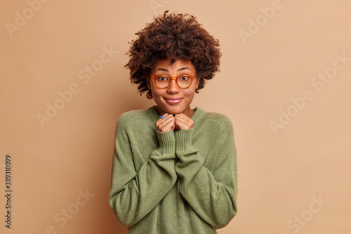 Pleased cheerful female student with Afro hair keeps hands under chin dressed in casual jumper wears glasses has charming smile on face isolated over beige background. Face expressions concept