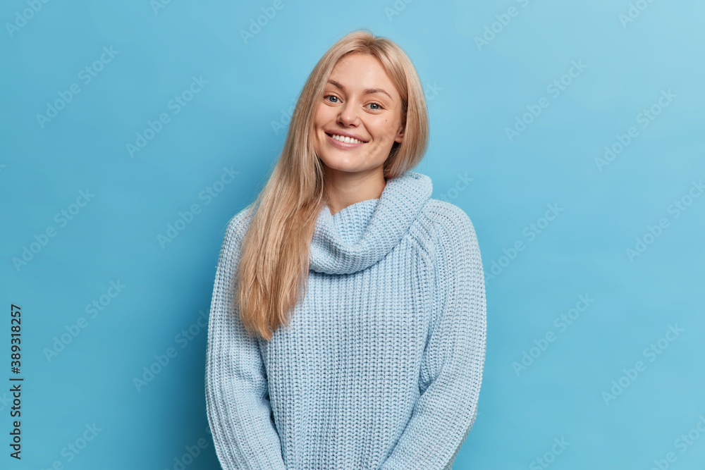 Pleased good looking woman with light hair smiles happily expresses good emotions glad to hear something pleasant wears knitted sweater models against blue background meets former classmate. Pleased good looking woman with light hair smiles happily expresses good emotions glad to hear something pleasant wears knitted sweater models against blue background meets former classmate.