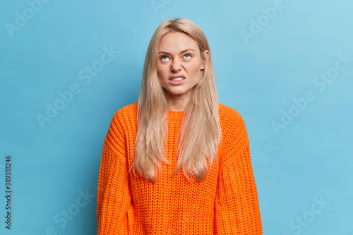 Slika na platnu Annoyed dissatisfied woman concentrated above smirks face and looks at something with aversion has long blonde hair dressed in knitted orange jumper isolated over blue wall