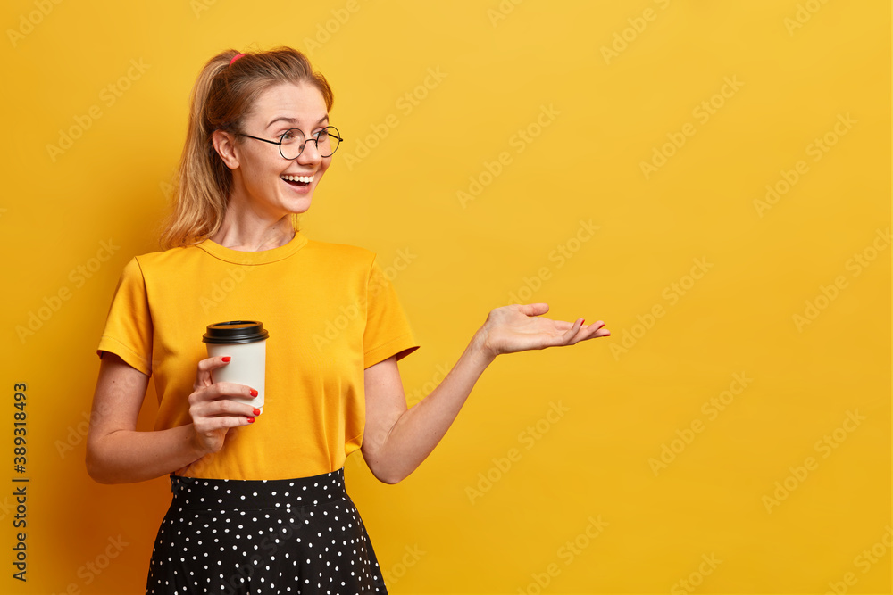 Sincere positive girl drinks aromatic coffee from disposable cup keeps hand raised over yellow empty background happy to see friend far away wears yellow t shirt and skirt being in good mood