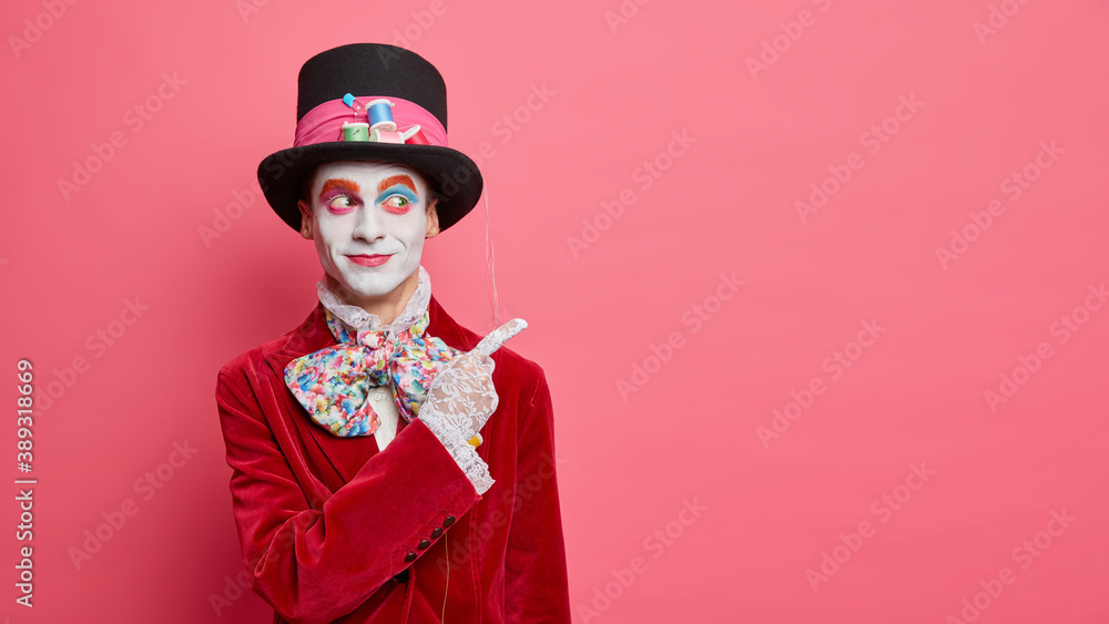 Street performer indicates at blank space and shows something wears bright makeup and costume isolated over pink studio background. Hatter from wonderland participates in halloween carnival. Street performer indicates at blank space and shows something wears bright makeup and costume isolated over pink studio background. Hatter from wonderland participates in halloween carnival.