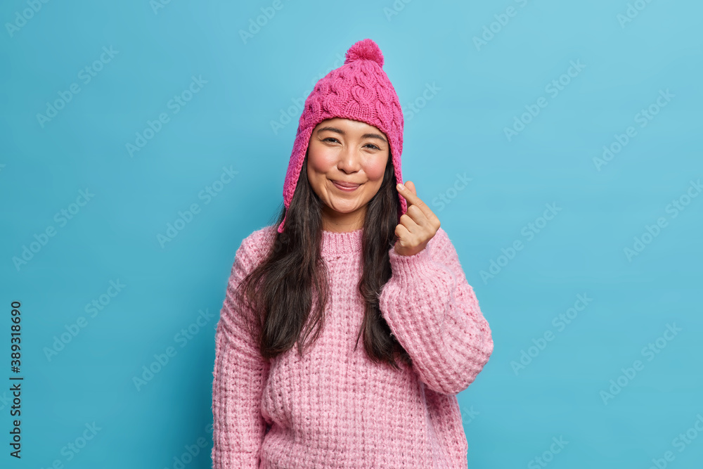 Pleased adorable romantic Asian woman makes korean like sign shapes mini heart at camera wears knitted hat and jumper poses against blue studio background. Body language concept. Love gesture Pleased adorable romantic Asian woman makes korean like sign shapes mini heart at camera wears knitted hat and jumper poses against blue studio background. Body language concept. Love gesture