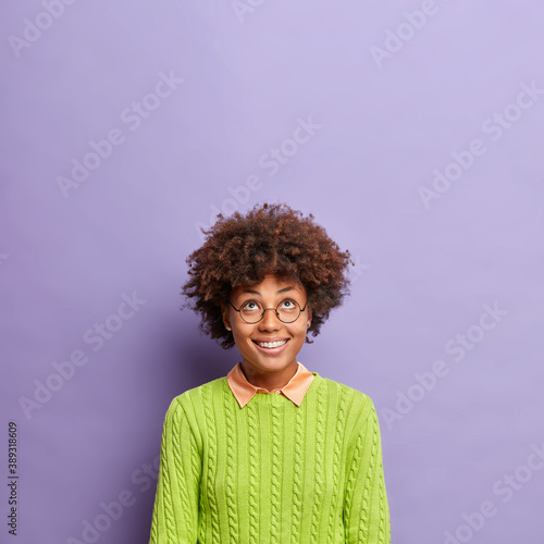 Vertical shot of happy curly haired young woman smiles broadly concentrated above sees something incredible looks upwards wears round spectacles and green jumper poses against purple background