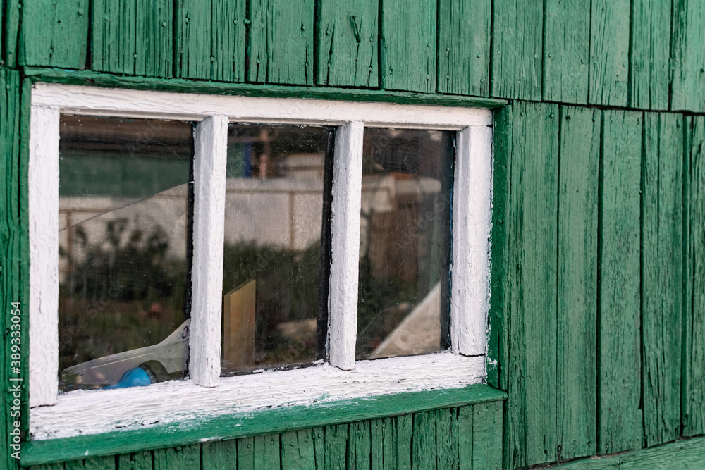 An old wooden small window with a frame of boards painted white and thin glass. On a battered wall of wooden planks with green paint. The wall of a dilapidated building with a rough surface An old wooden small window with a frame of boards painted white and thin glass. On a battered wall of wooden planks with green paint. The wall of a dilapidated building with a rough surface