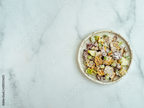 Waldorf salad with copy space. American fruit and nut salad with apples, celery, grapes, chicken meat, dressed mayonnaise. Craft plate with ready-to-eat waldorf salad on white marble. Top view