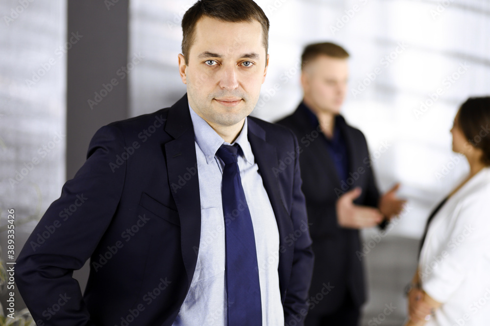 Portrait of a self-confident middle aged businessman in a blue suit, standing in a modern office with his colleagues at the background. Concept of business success Portrait of a self-confident middle aged businessman in a blue suit, standing in a modern office with his colleagues at the background. Concept of business success