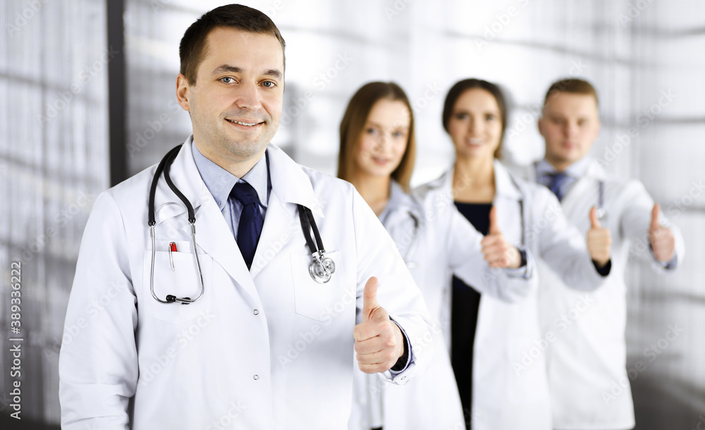 Group of professional doctors are standing as a team with thumbs up in a hospital office, ready to help their patients. Medical help, insurance in health care and medicine concept