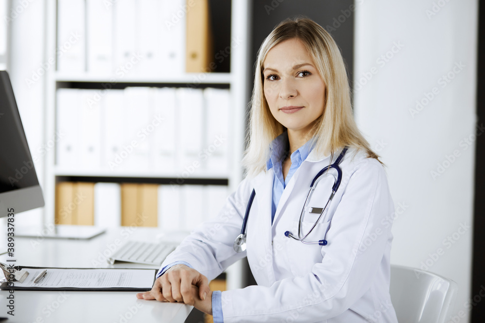 Friendly female doctor sitting in clinic at her working place. Portrait of cheerful smiling physician. Perfect medical service in hospital. Medicine and healthcare concept Friendly female doctor sitting in clinic at her working place. Portrait of cheerful smiling physician. Perfect medical service in hospital. Medicine and healthcare concept