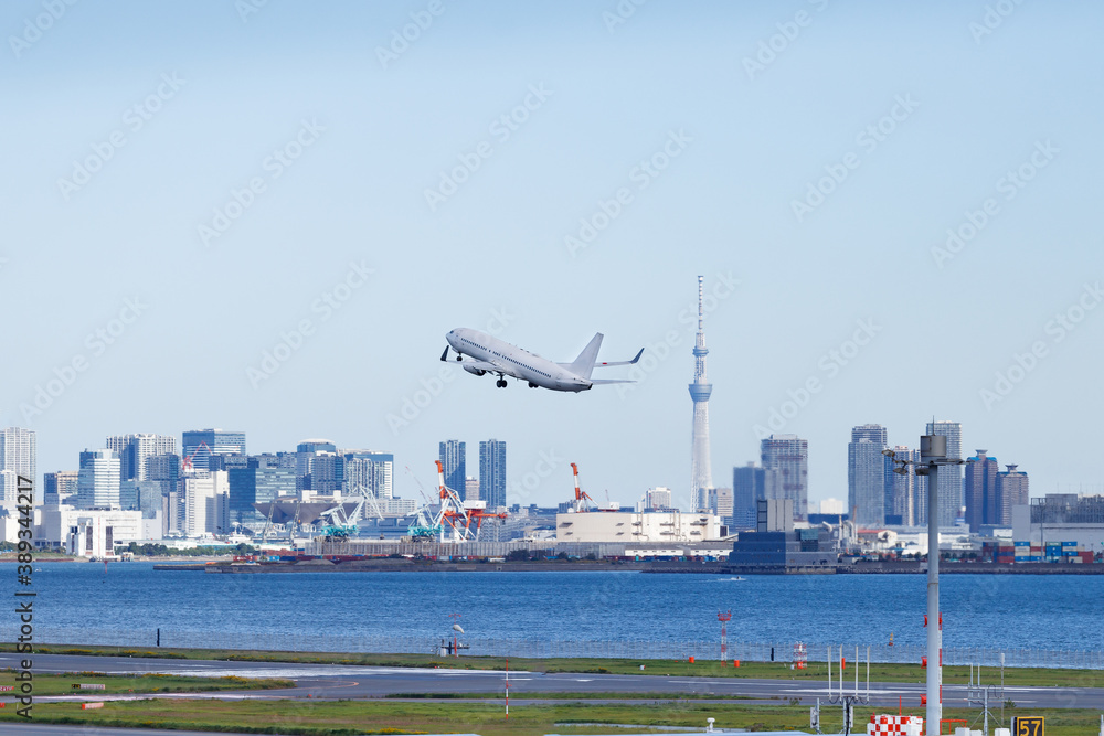 青空を背景に羽田空港を離陸する飛行機と東京湾岸のビル群とスカイツリー Stock Photo Adobe Stock