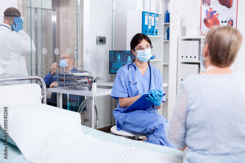 Medical staff wearing protective mask against coronavirus pandemic and talking with senior patient during consultation in medical examination room. Practitioner physician appointment