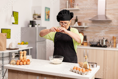 Fototapeta Naklejka Na Ścianę i Meble -  A woman prepares a dough for baking cracking eggs in home kitchen. Elderly pastry chef cracking egg on glass bowl for cake recipe in kitchen, mixing by hand, kneading ingredients prreparing homemade