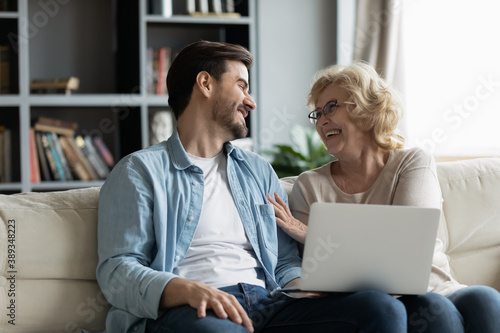 Happy middle aged mature woman in eyewear laughing at funny comedian film or family vacation photos with joyful grown up son, enjoying using computer applications together at home, technology concept.